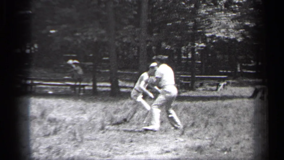 Two adult men bend and reach during the casual kickball-style game, with trees and sideline figures visible behind them in Cleveland footage.