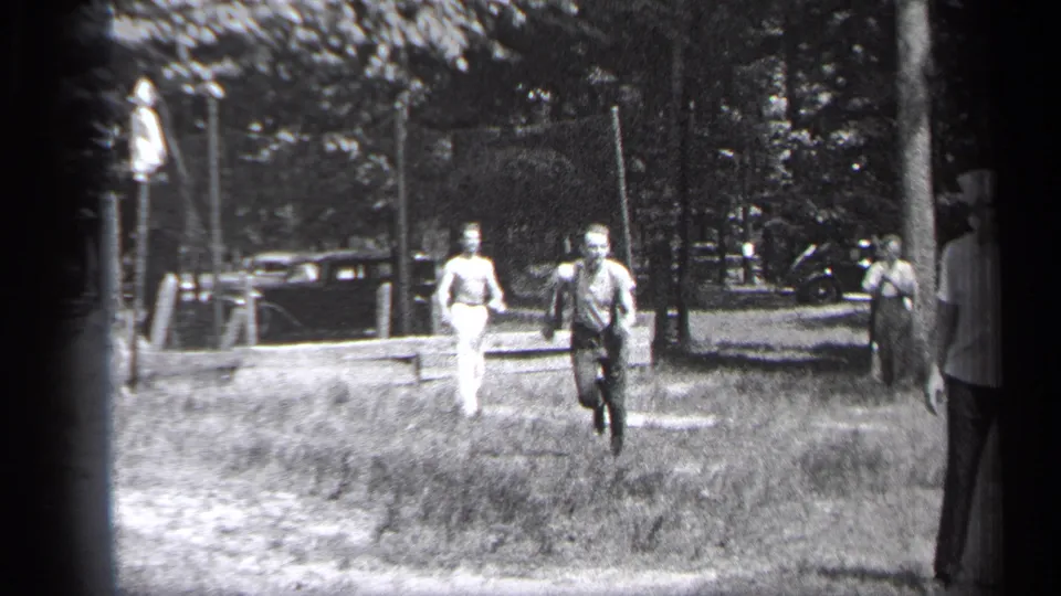Two adult players run toward the camera in 1937 Cleveland home-movie footage, with spectators, trees, and a bench or low barrier behind them.