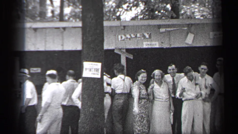 1937 Cleveland sidewalk gathering with adults clustered near a tree and storefront sign, captured in soft black-and-white 8mm framing.