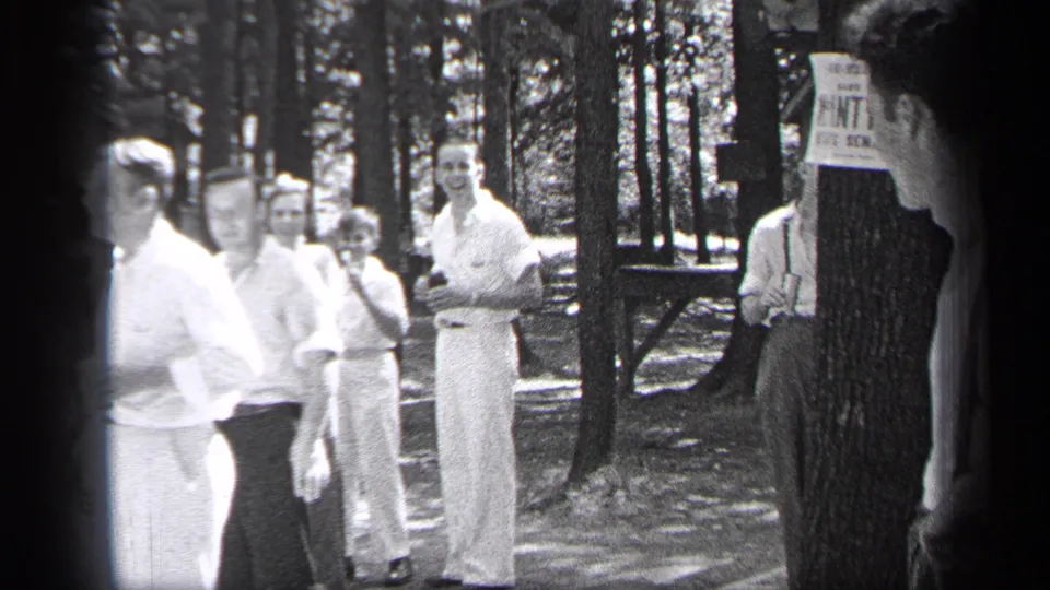 1937 Cleveland home-movie frame showing a man in a light suit near onlookers, trees, and a posted notice at the edge of a sidewalk gathering.