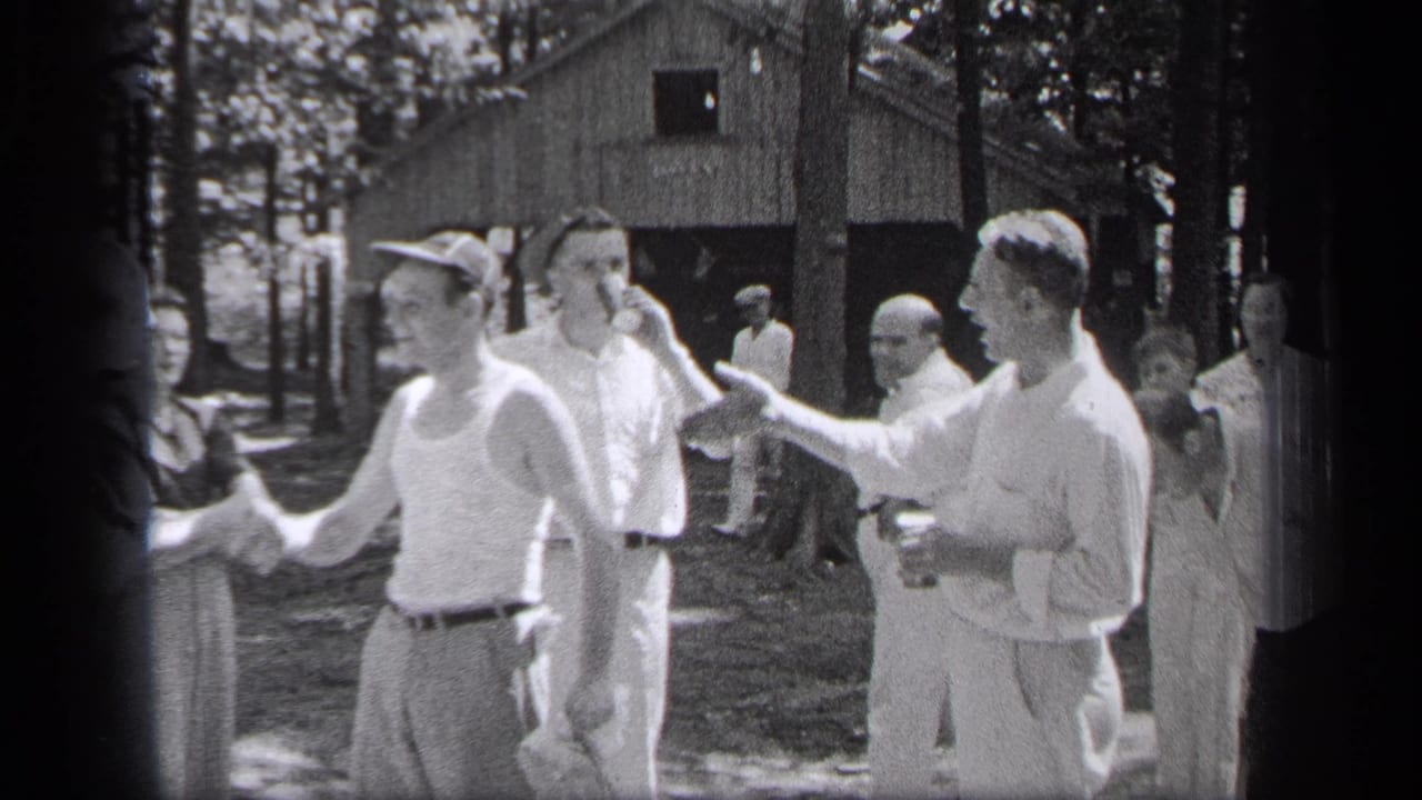 Men in summer clothing shake hands near trees and a rustic cabin.