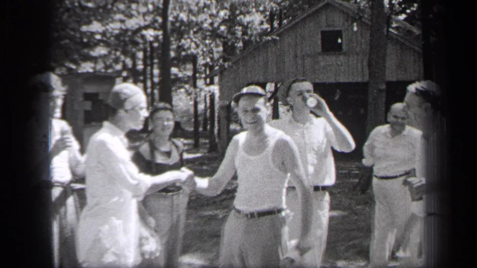 A man in a cap shakes hands in a wooded Cleveland setting while other men stand nearby, including one drinking from a can.