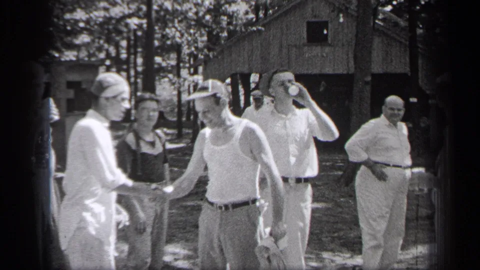 Two men shake hands in front of a rustic building while several adult men in light clothing stand around in the wooded area.