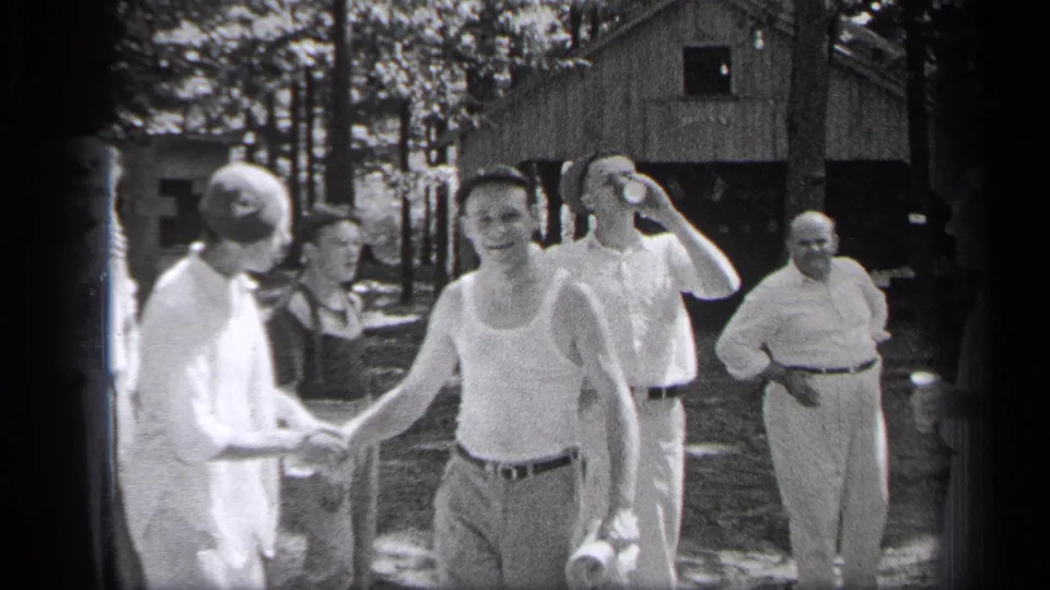 In a black-and-white 1937 Cleveland frame, a man in a tank top faces the camera while others continue greeting each other outdoors.