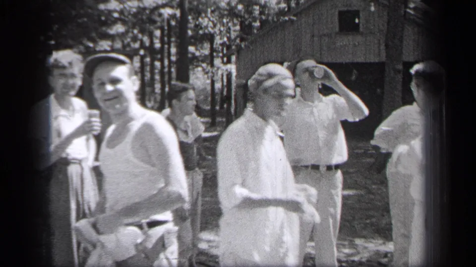 1937 Cleveland outdoor home-movie frame with adults in light clothing socializing beside trees and a cabin.