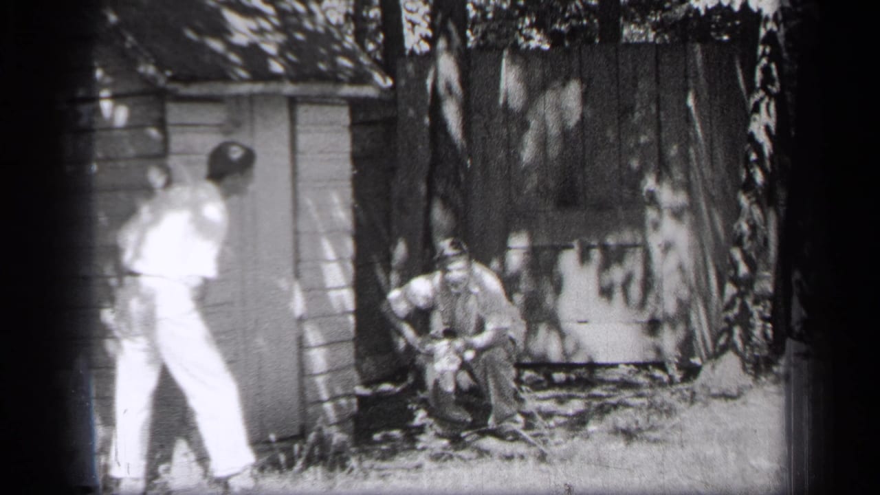 Two men near a wooden shed, one standing and one crouching in a shaded yard.