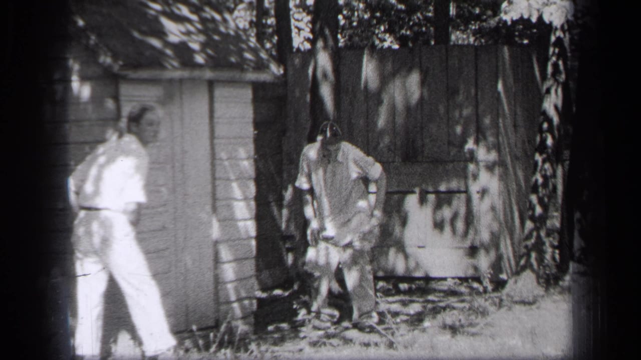 A man in light clothing stands near a shed while another man stands by yard debris.