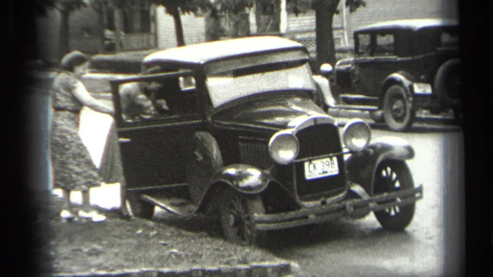 1938 Cleveland home-movie frame of a woman in a patterned dress assisting someone into a black sedan on a residential street.
