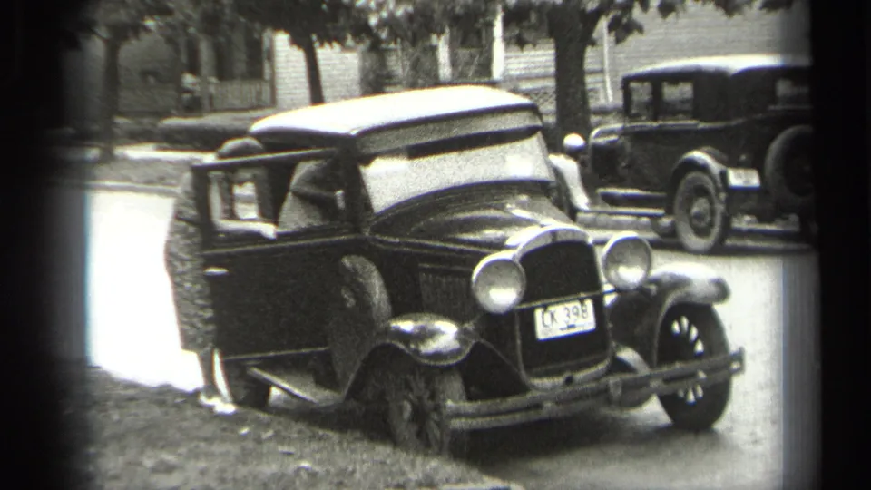 The black sedan remains parked at the curb in Cleveland, with one adult partly visible by the open door and another vintage car behind it.