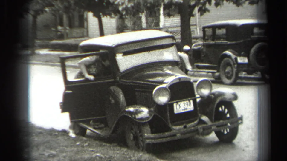 An adult leans into the open door of the 1938 Cleveland street scene, with the sedan front, license plate, and background car visible.
