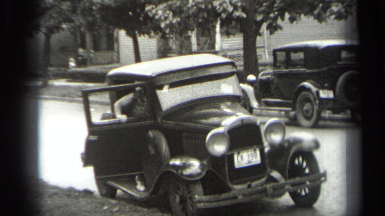 Seated driver in a dark sedan with the door open and another car behind.