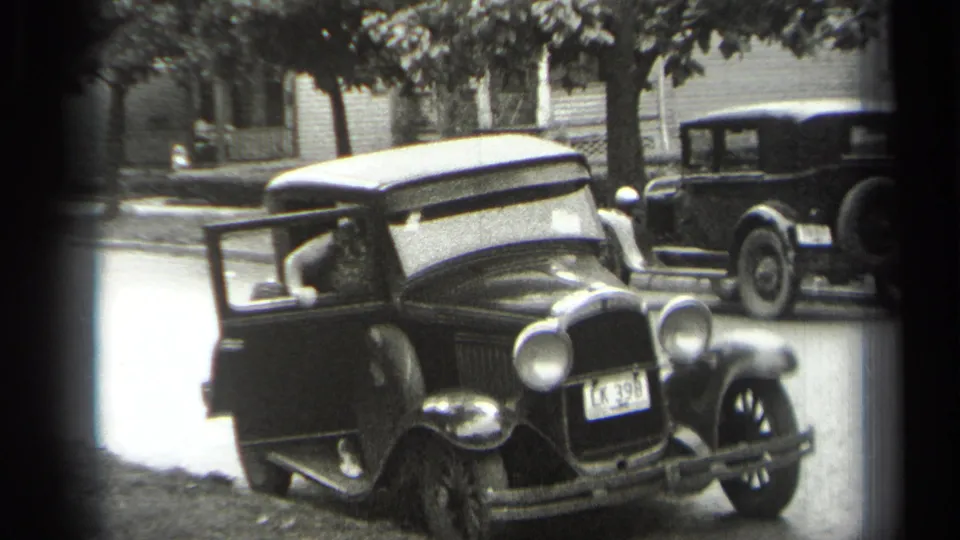 Cleveland 1938 frame with a man visible in the driver's seat of a dark sedan, its door open beside a curbside street.