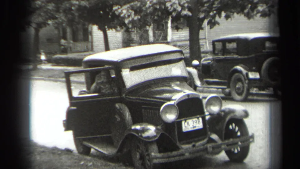 Cleveland 1938 home-movie still showing the driver inside a dark sedan with its door open, front plate visible, and a parked car in back.