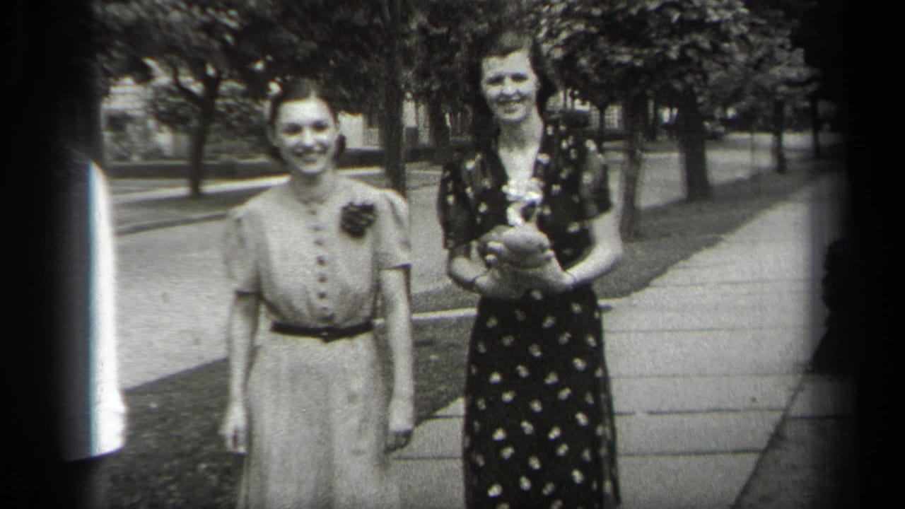 Two women stand smiling on a tree-lined sidewalk, one in a light dress and one in a dark patterned dress.