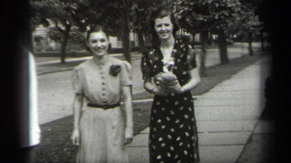 In 1938 Cleveland home-movie footage, two young women pose on a sidewalk in vintage dresses, with trees and a street behind them.