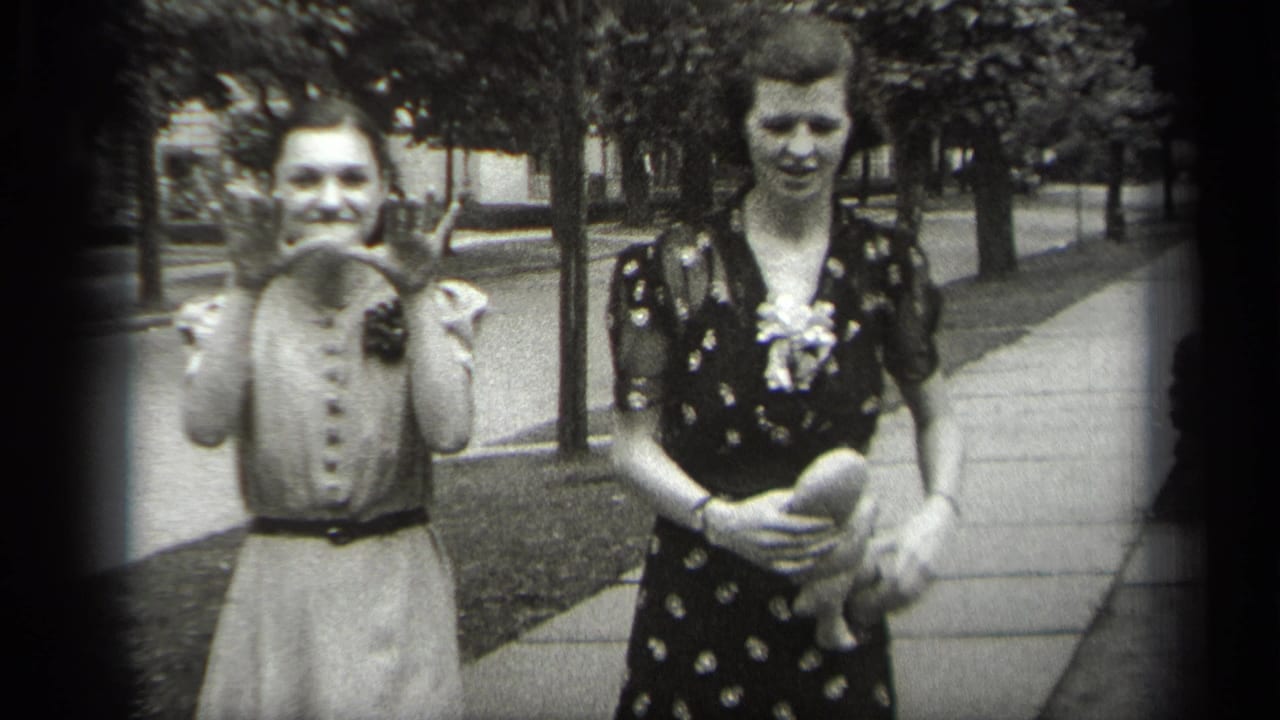 Two women face the camera on a sidewalk, one raising her hands while the other holds a small doll.