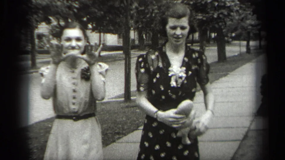 The two women remain on the Cleveland sidewalk as one waves or gestures toward the camera and the other looks down while holding a doll.