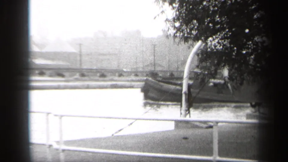 1938 Cleveland home-movie frame showing a tugboat approaching along a river, with foreground railing, tree branches, and a hazy city backdrop.
