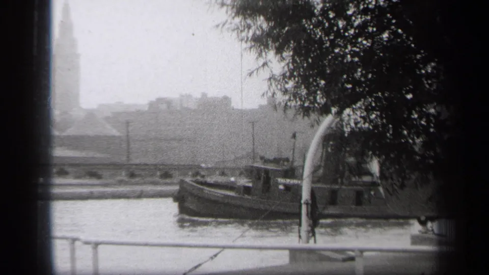 Black-and-white 8mm frame of a tugboat on a Cleveland river, seen through dark frame edges with trees and a faint skyline behind it.