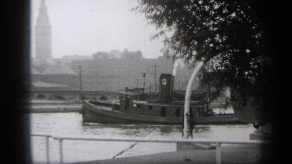 1938 Cleveland frame showing the tugboat in side profile on the river, with a distant clock tower, low buildings, trees, and railings visible.