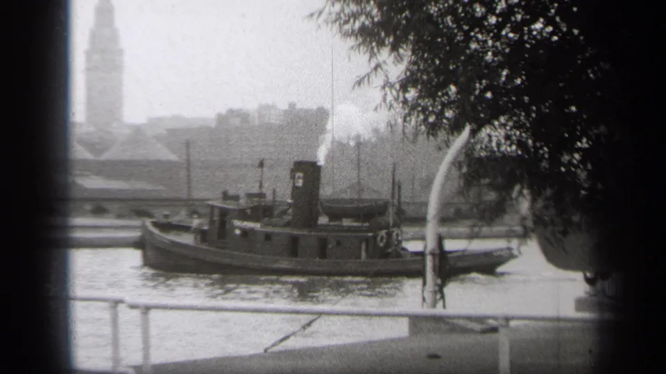 Black-and-white 8mm frame of a tugboat centered on a Cleveland river, with foreground railings, tree branches, and a hazy clock tower beyond.