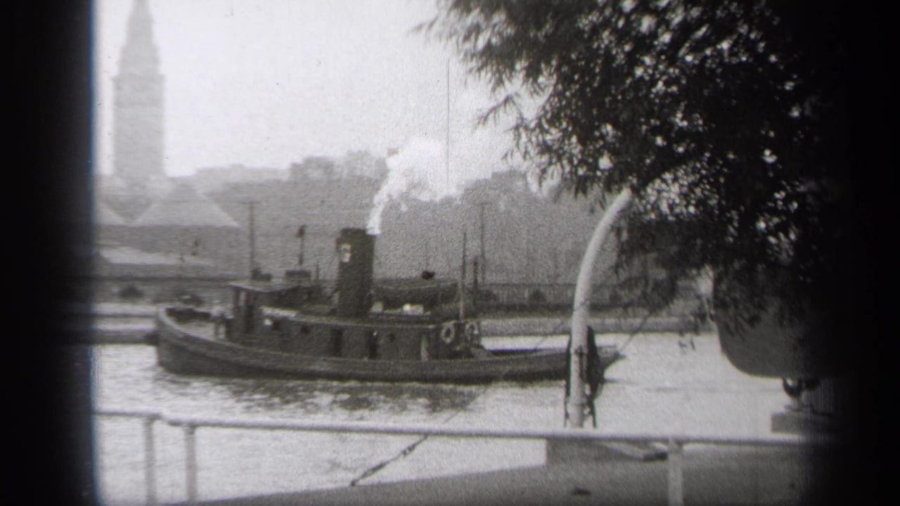 1938 Cleveland waterfront view with a tugboat moving across the river, smoke from its stack, railing in front, trees at right, and a tower in the distance.