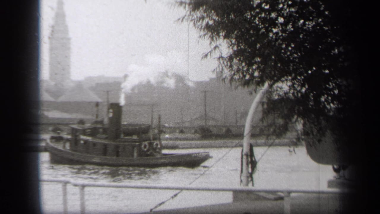 Tugboat centered on the river beneath tree branches, with smoke and a distant tower.