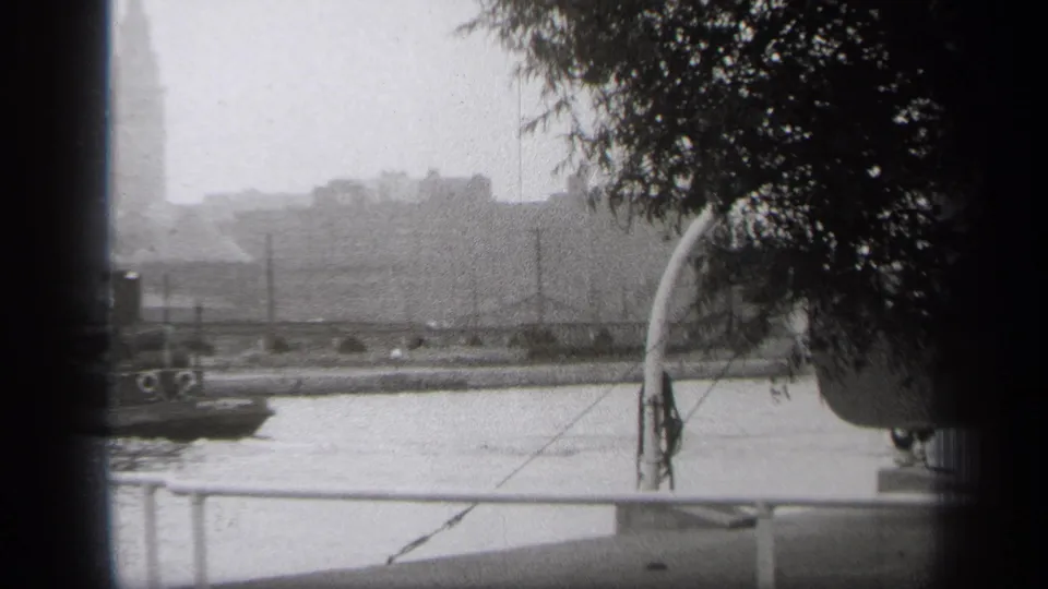 1938 Cleveland home-movie frame showing a riverfront view with foreground railing, trees, distant skyline, and a tall tower in haze.