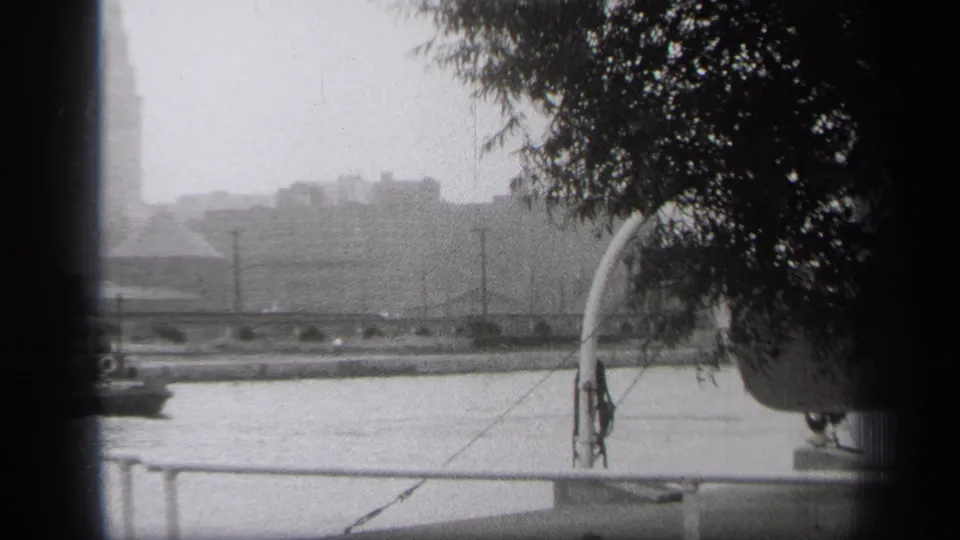 1938 Cleveland river scene with railing in front, distant buildings and tower beyond the water, and tree cover along the right edge.