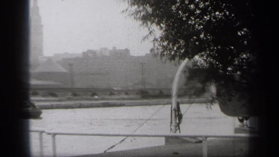 Cleveland riverfront home-movie frame from 1938, showing water, a foreground rail, overhanging branches, and a hazy tower in the distance.
