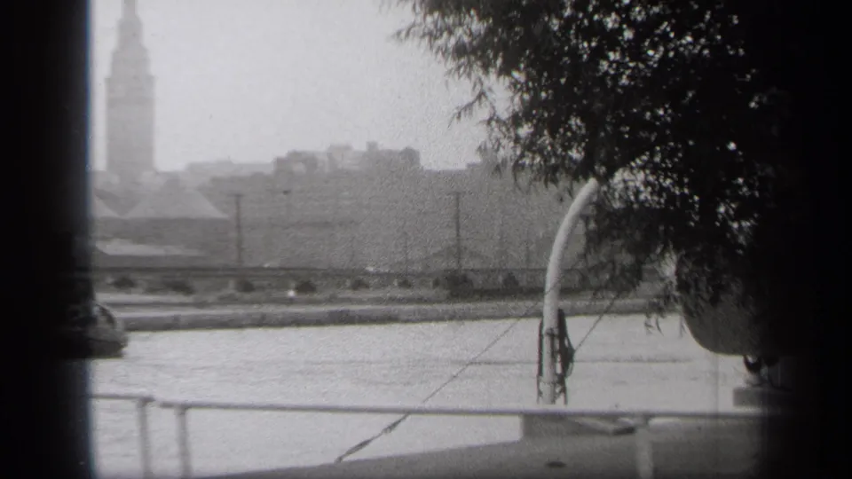1938 Cleveland 8mm frame looking across the river toward a distant urban skyline, with railing and tree branches in the foreground.
