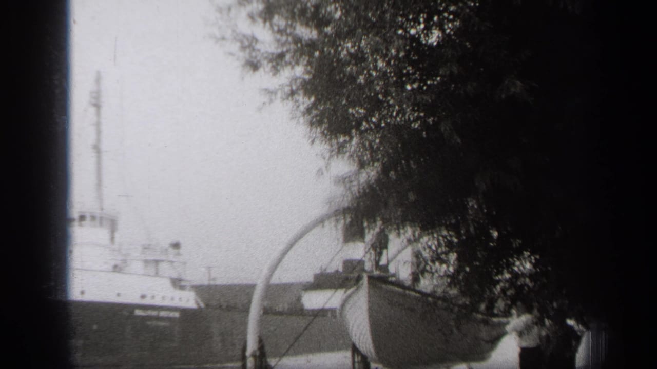 1938 Cleveland home-movie frame showing a cargo ship on the water, viewed past trees and a foreground boat on a trailer.