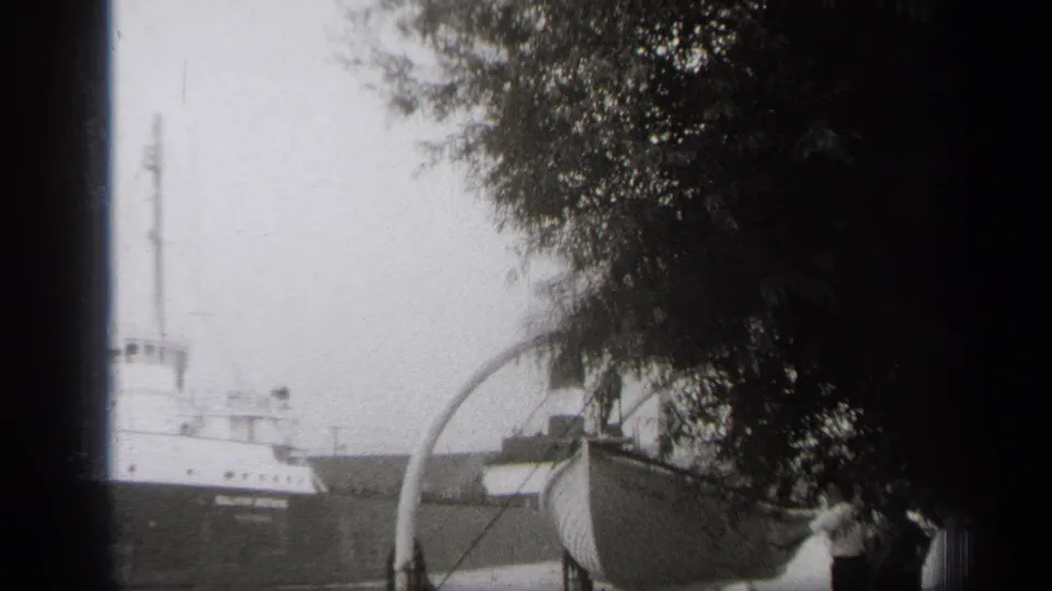 1938 Cleveland home-movie frame showing a cargo ship on the water, viewed past trees and a foreground boat on a trailer.