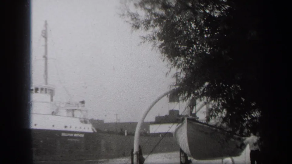 Cleveland 8mm frame showing a passing cargo ship behind a foreground boat, tree cover, and a partially visible person near the shore.