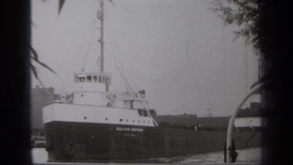 1938 Cleveland home-movie frame showing the Sullivan Brothers vessel stationary in calm water, partly framed by dark leaves.