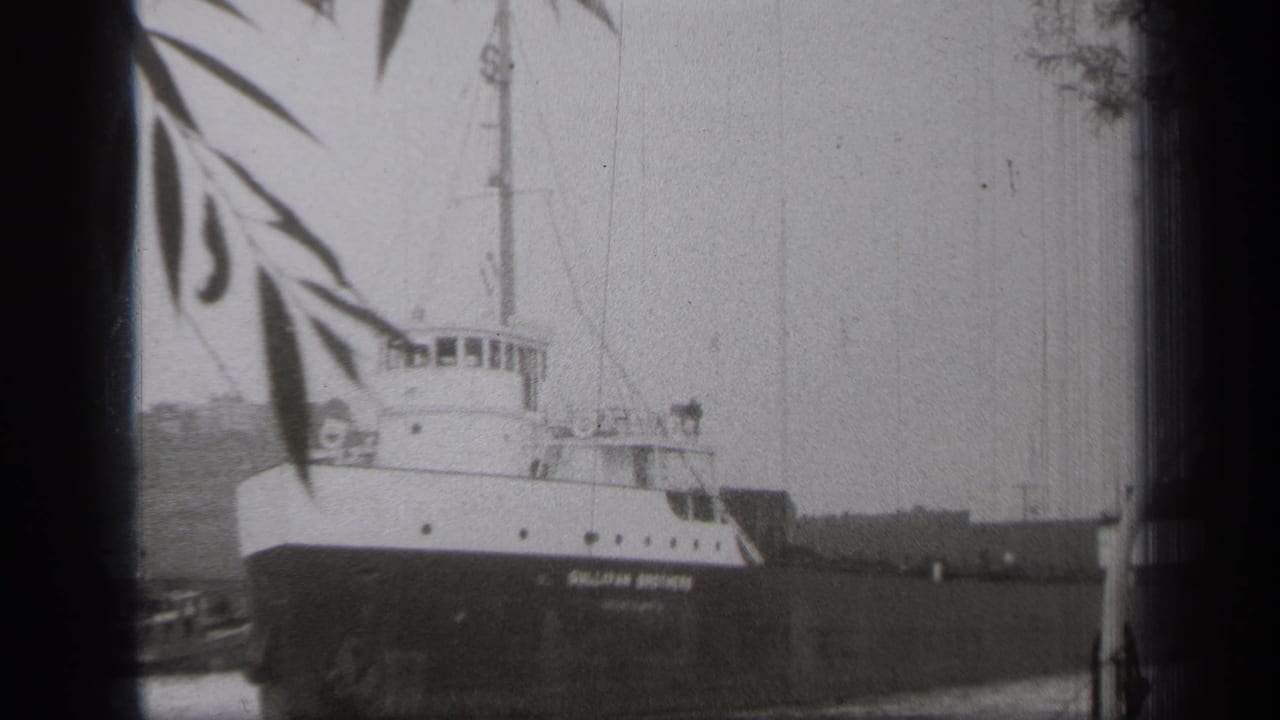 Stationary ship on calm water with mast and dockside details visible.