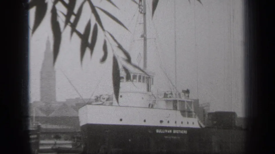 1938 Cleveland harbor view of the SULLIVAN BROTHERS vessel framed by dark tree leaves, with a tall waterfront building behind it.