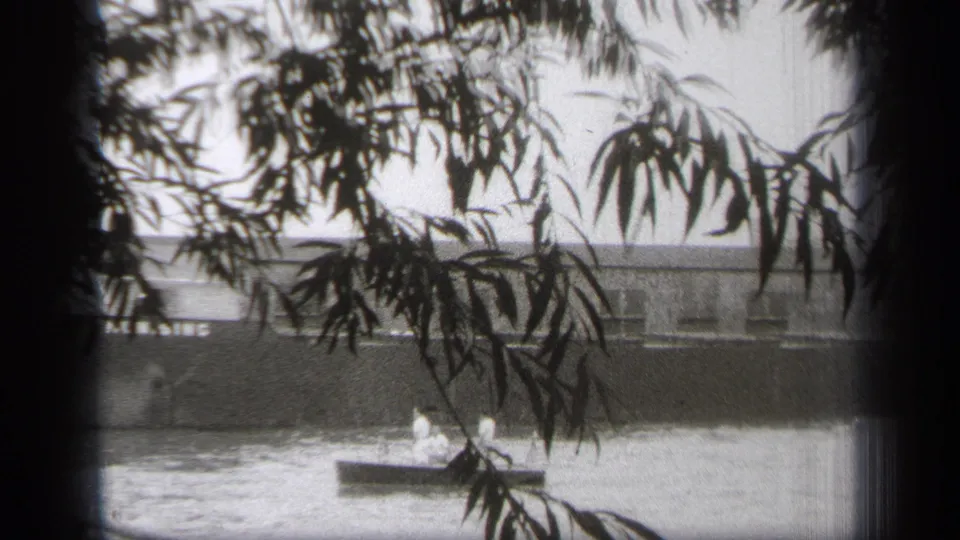 Black-and-white 8mm frame of a small boat on the Cleveland waterfront, seen through branches with a long building across the water.