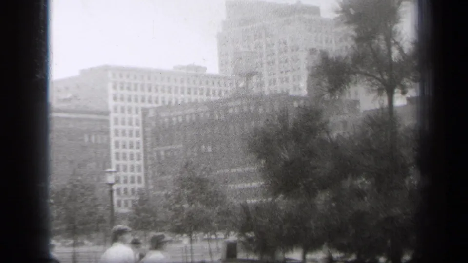 1938 Cleveland cityscape view showing multi-story buildings, trees, a street, and distant pedestrians in soft black-and-white 8mm footage.