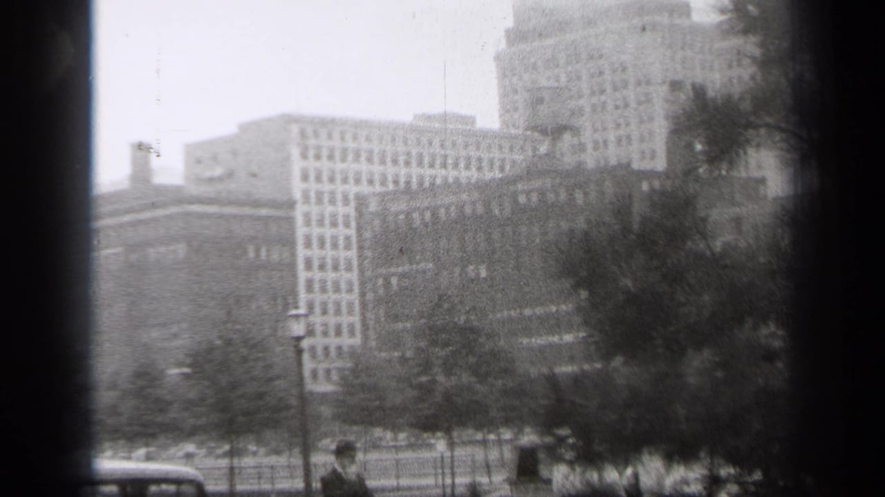 1938 Cleveland street-level view across park trees toward downtown buildings and a tall tower, with parked cars and pedestrians at the bottom of frame.