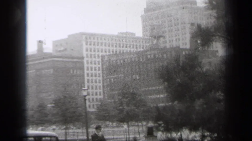 1938 Cleveland street-level view across park trees toward downtown buildings and a tall tower, with parked cars and pedestrians at the bottom of frame.