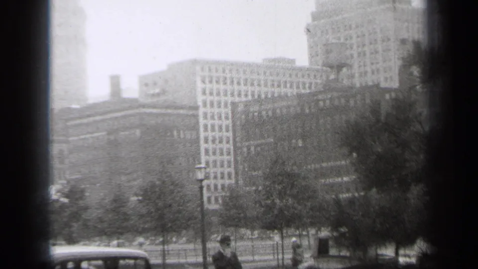 The pan continues across Cleveland buildings behind a tree-lined park area, with a period automobile and small pedestrians visible in the foreground.