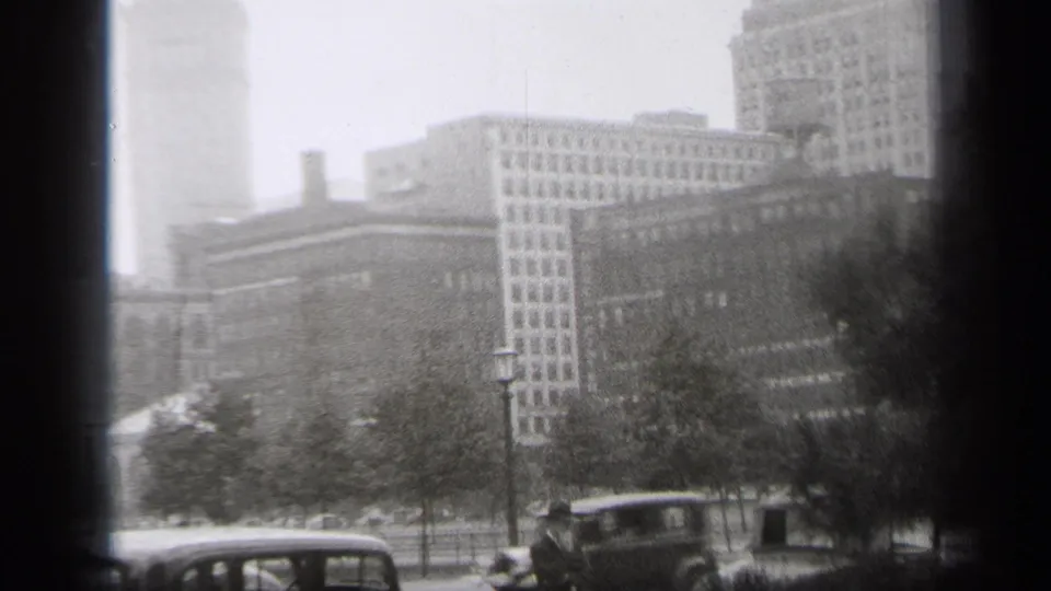 Soft 8mm frame showing Cleveland architecture beyond park trees, with multiple 1930s automobiles crossing or parked along the street foreground.