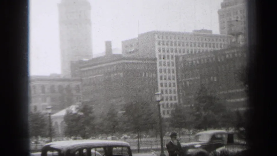 Late pan frame from 1938 Cleveland, showing a dark car, a pedestrian, park trees, lampposts, and large downtown buildings behind them.