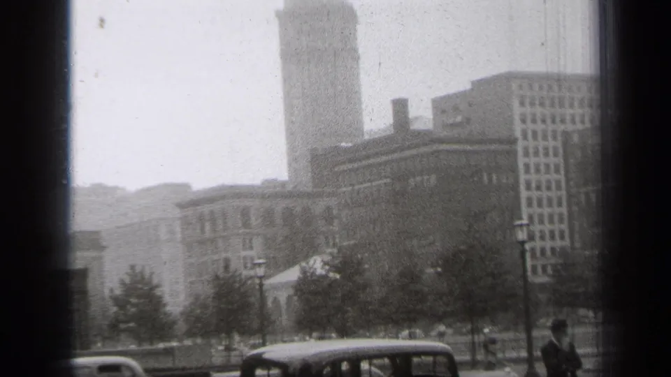1938 Cleveland street scene showing period cars, trees, lampposts, a tall tower, and a man in dark clothing near the right edge.