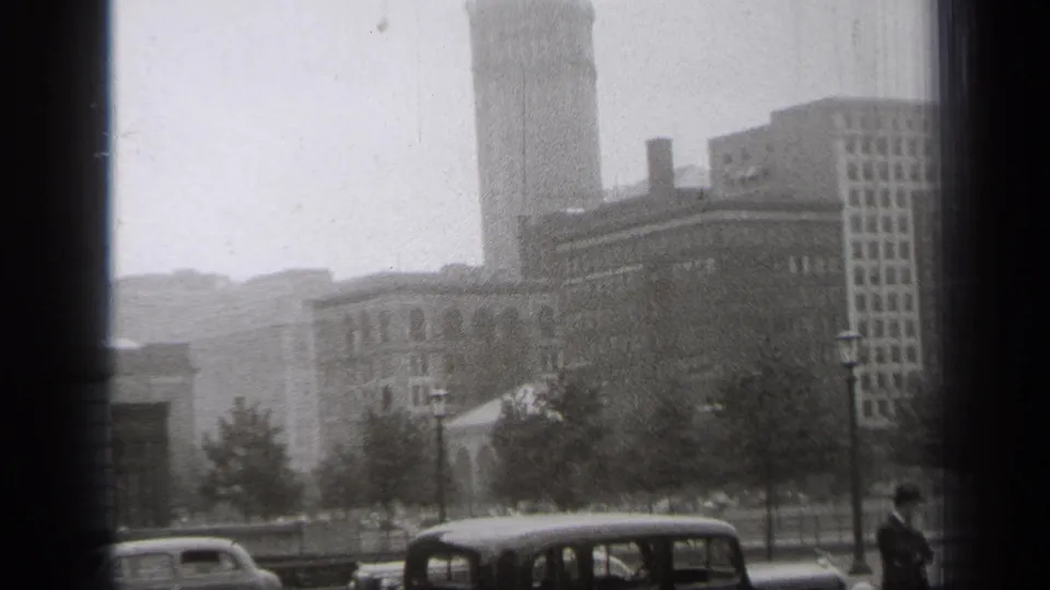 1938 Cleveland view with classic automobiles, a row of trees, lampposts, downtown buildings, and a hazy tower behind a dark border.