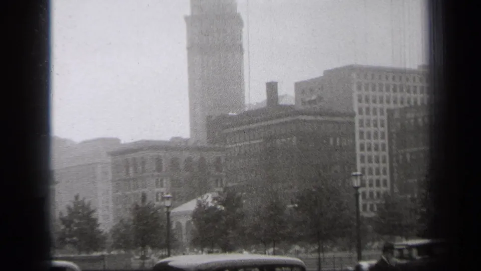 1938 Cleveland street view with parked cars, a suited pedestrian near the edge, trees, lampposts, and a tall tower in the background.
