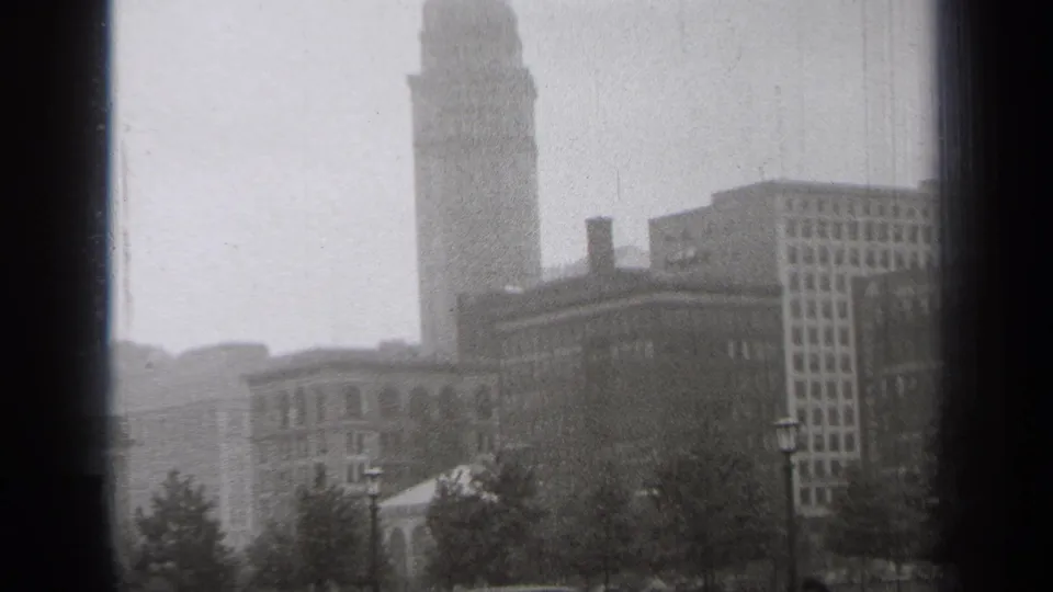 1938 Cleveland skyline view with a tall ornate tower, surrounding buildings, street lamps, trees, and soft fog.