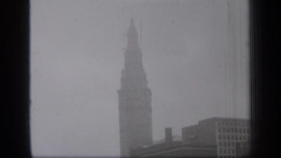 1938 Cleveland home-movie frame showing a tall spired building and nearby rooftops through a dark window edge.