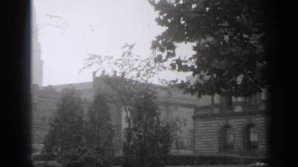 1938 Cleveland frame with leafy trees, masonry facades, rooftops, and a dark window mullion in soft black-and-white film.